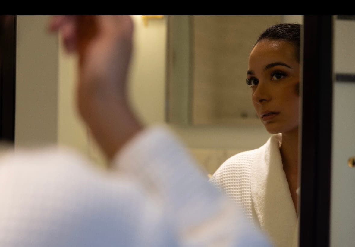 An editorial portrait framed through a doorway, a woman in a pale knit cardigan looking toward the camera with a blurred arm crossing the foreground.
